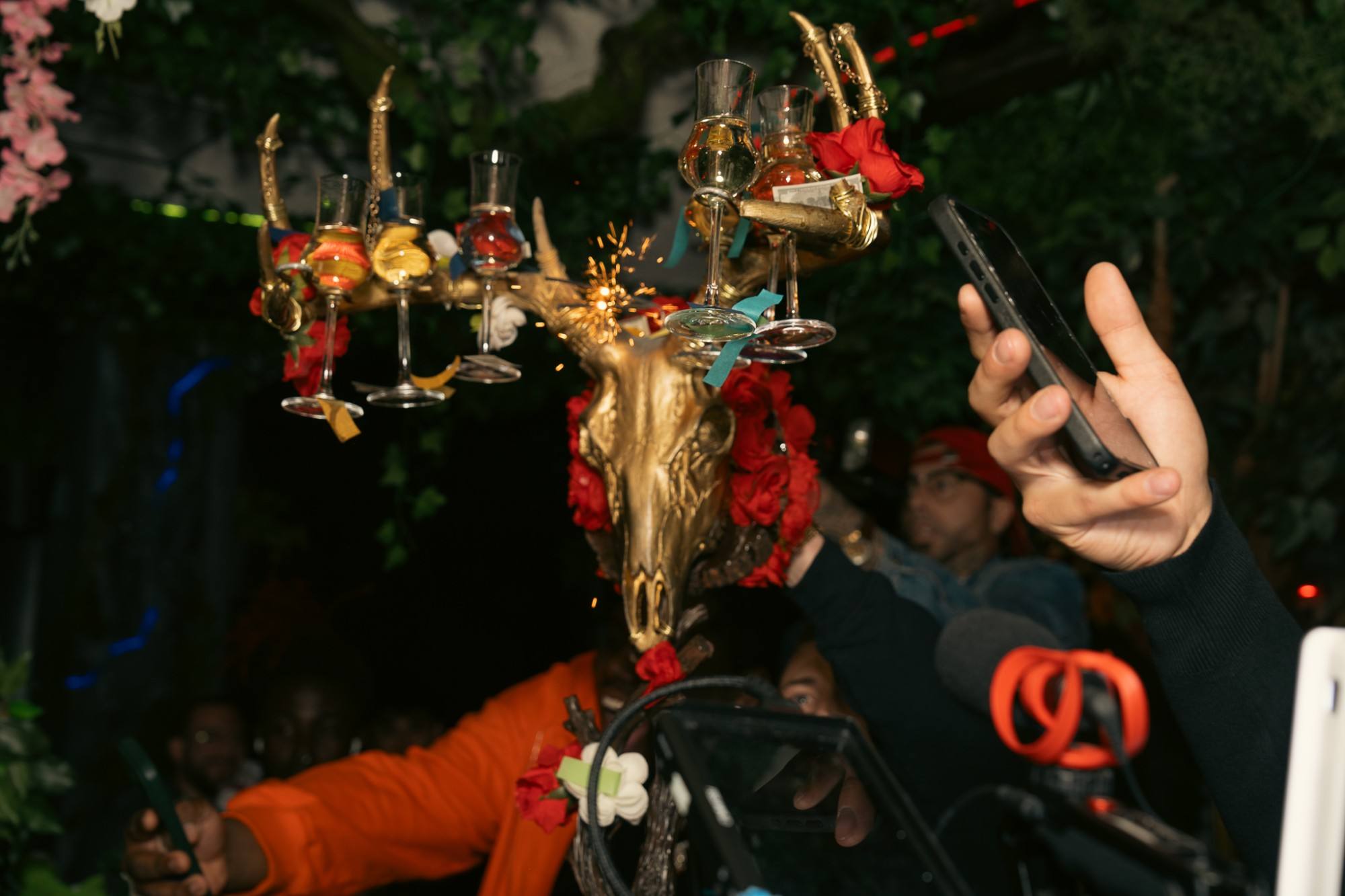 The Money Shot: a gold horned skull hung with red roses and stemmed shot glasses, a sparkler throwing sparks, as guests hold up their phones to film it.